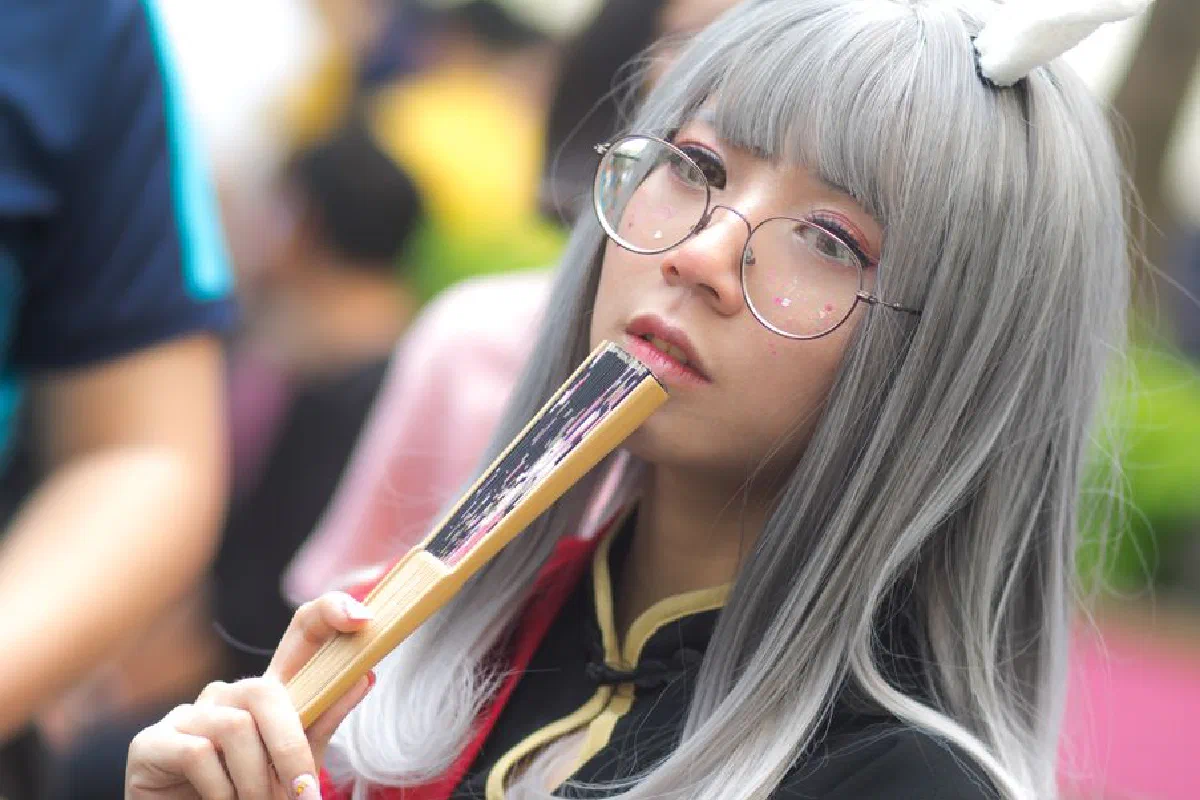 A stylish young woman in cosplay, wearing glasses and holding a fan, posing outdoors.