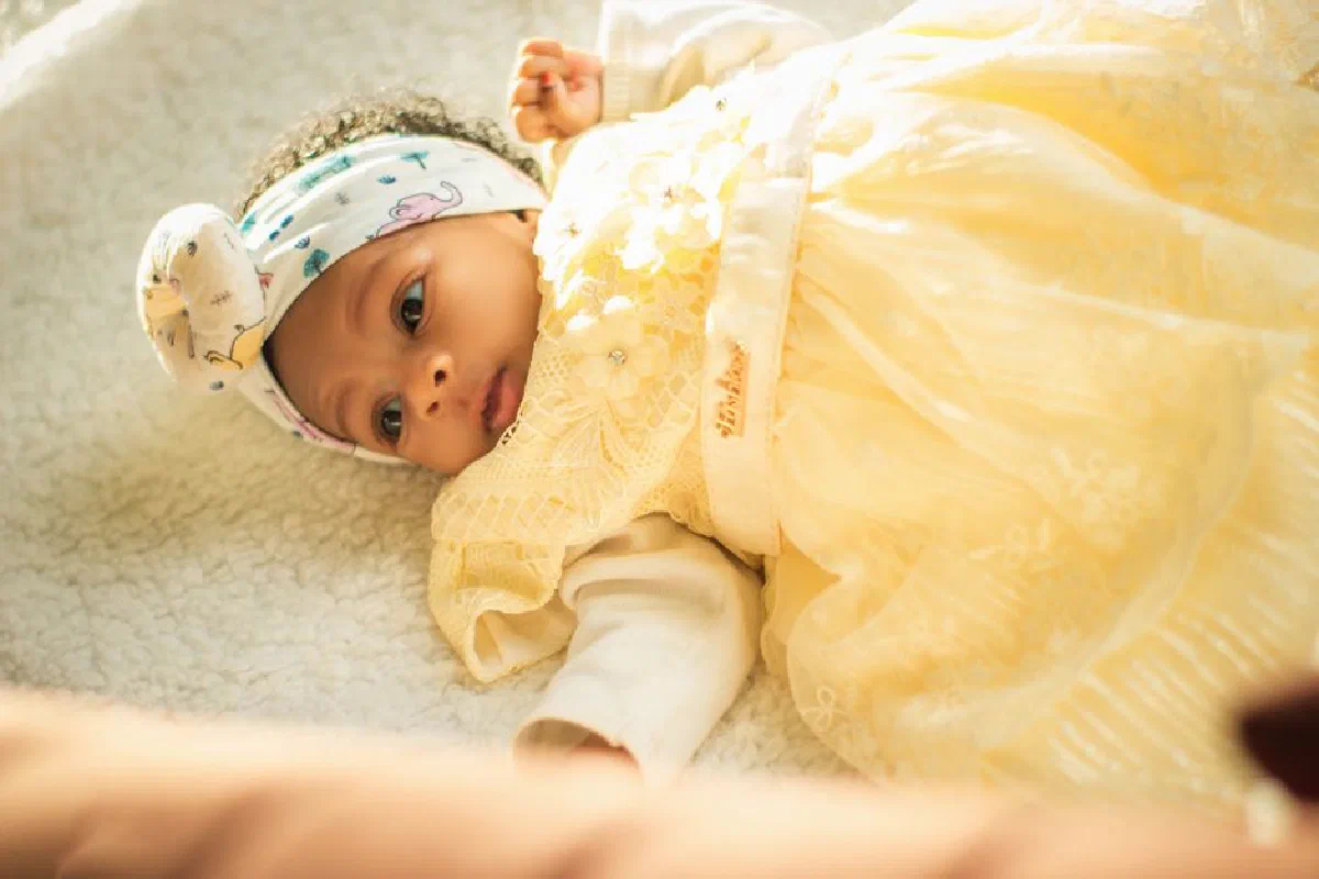Cute baby wearing a yellow lace dress with a patterned headband lying on a soft blanket.