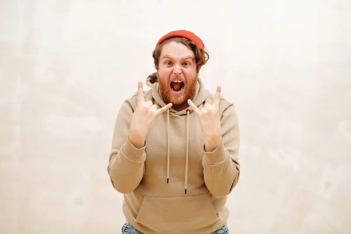 A bearded man in a beige hoodie enthusiastically shows a rock hand sign indoors.