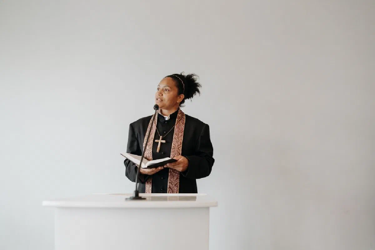 A female pastor delivers a sermon at a church podium, holding a Bible and wearing a cross.