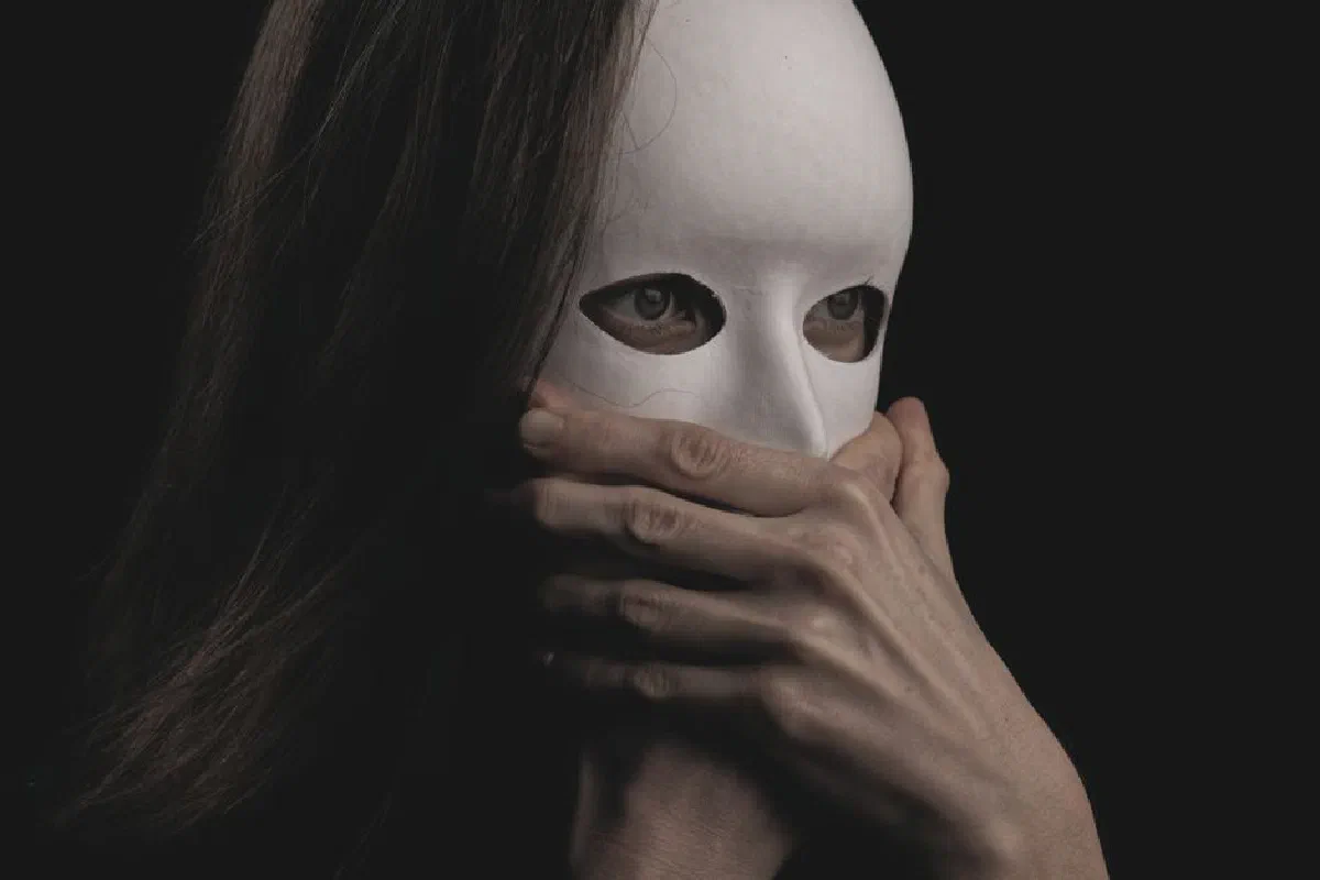 Close-up of a woman in a white mask, hands covering mouth, evoking mystery and drama.
