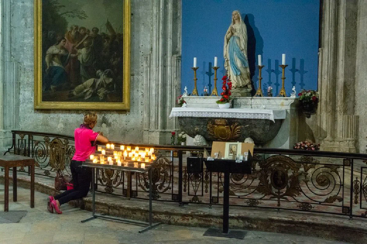 A woman kneels by lit candles, praying in a serene church setting.