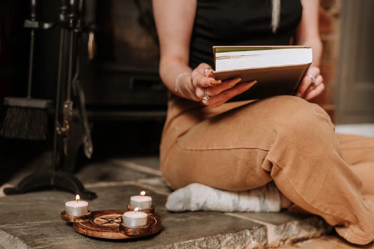 Crop anonymous female with book in hands near pentagram candle holder on floor