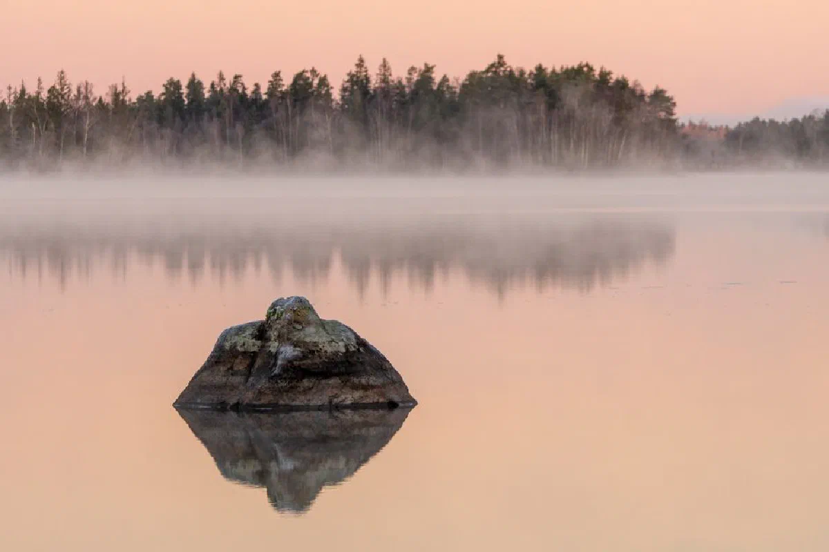 Tranquil lake scene at sunrise with mist and trees reflected in calm waters.