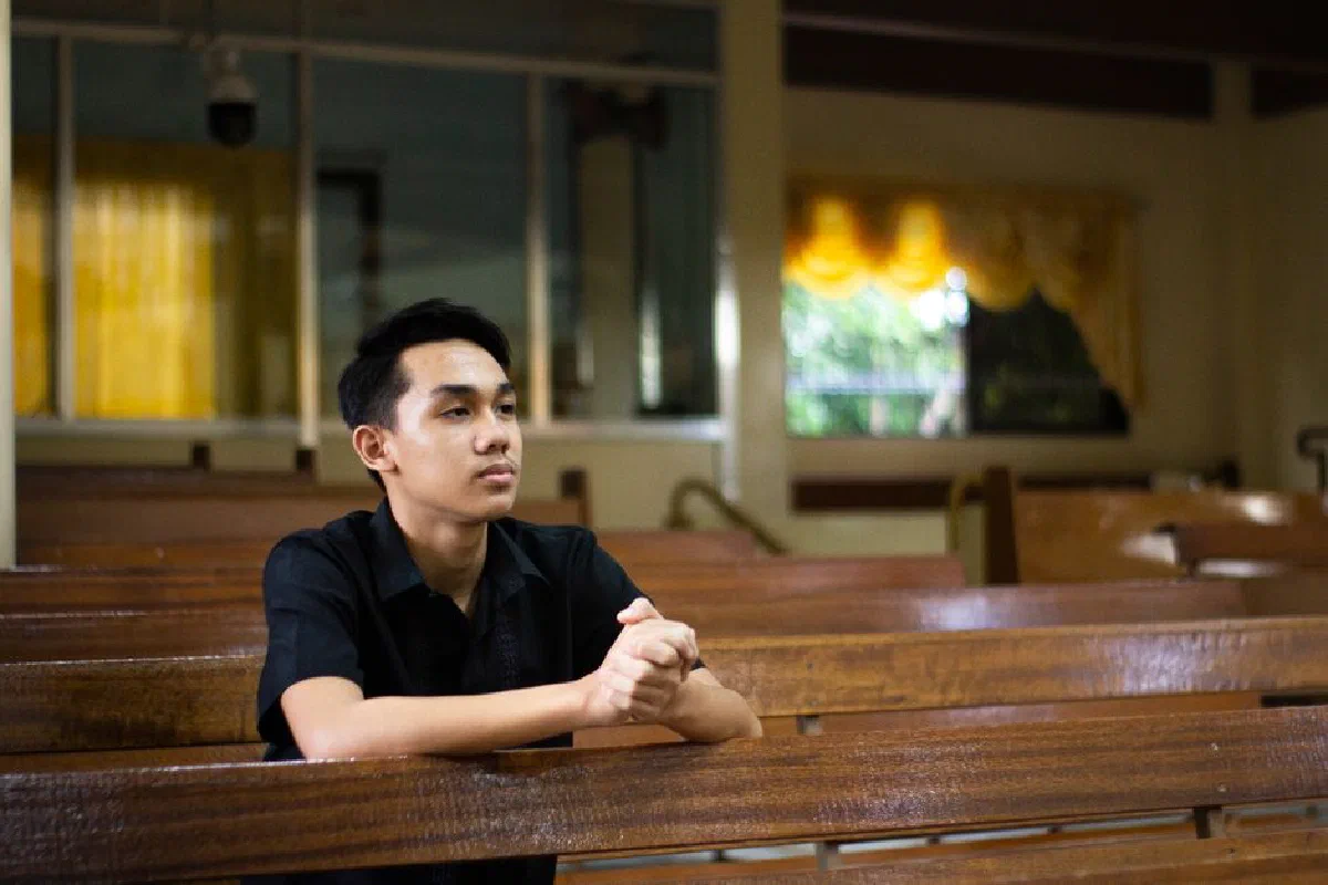 A young man in a church, sitting in a pew, clasping hands in prayer.