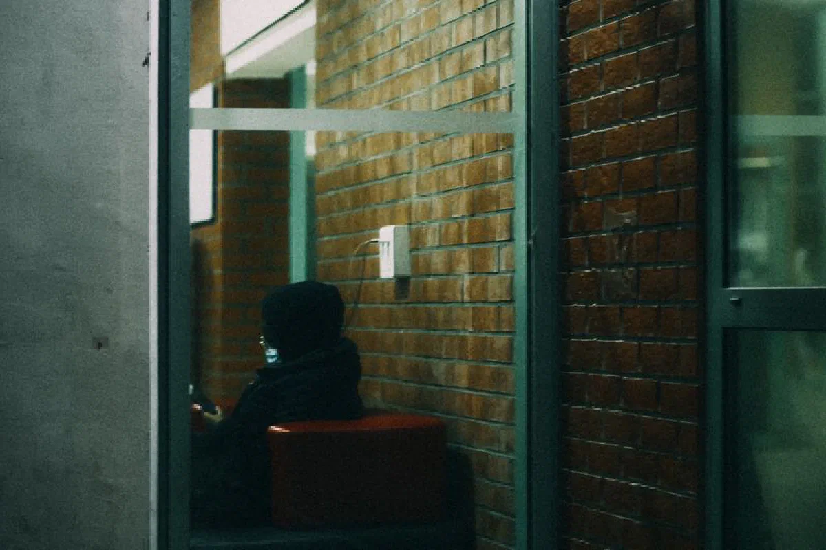 Person in a hoodie sitting indoors against a brick wall, using a phone.