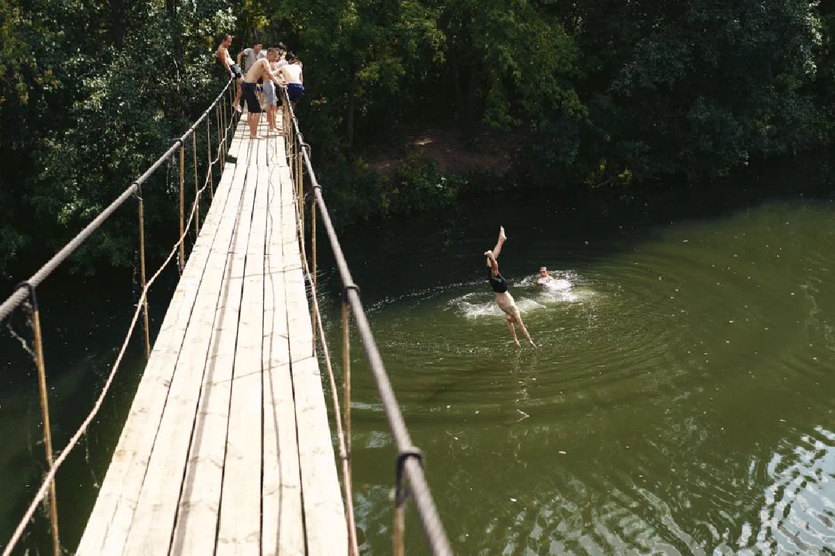 Friends dive into a river from a footbridge, enjoying a sunny summer day.