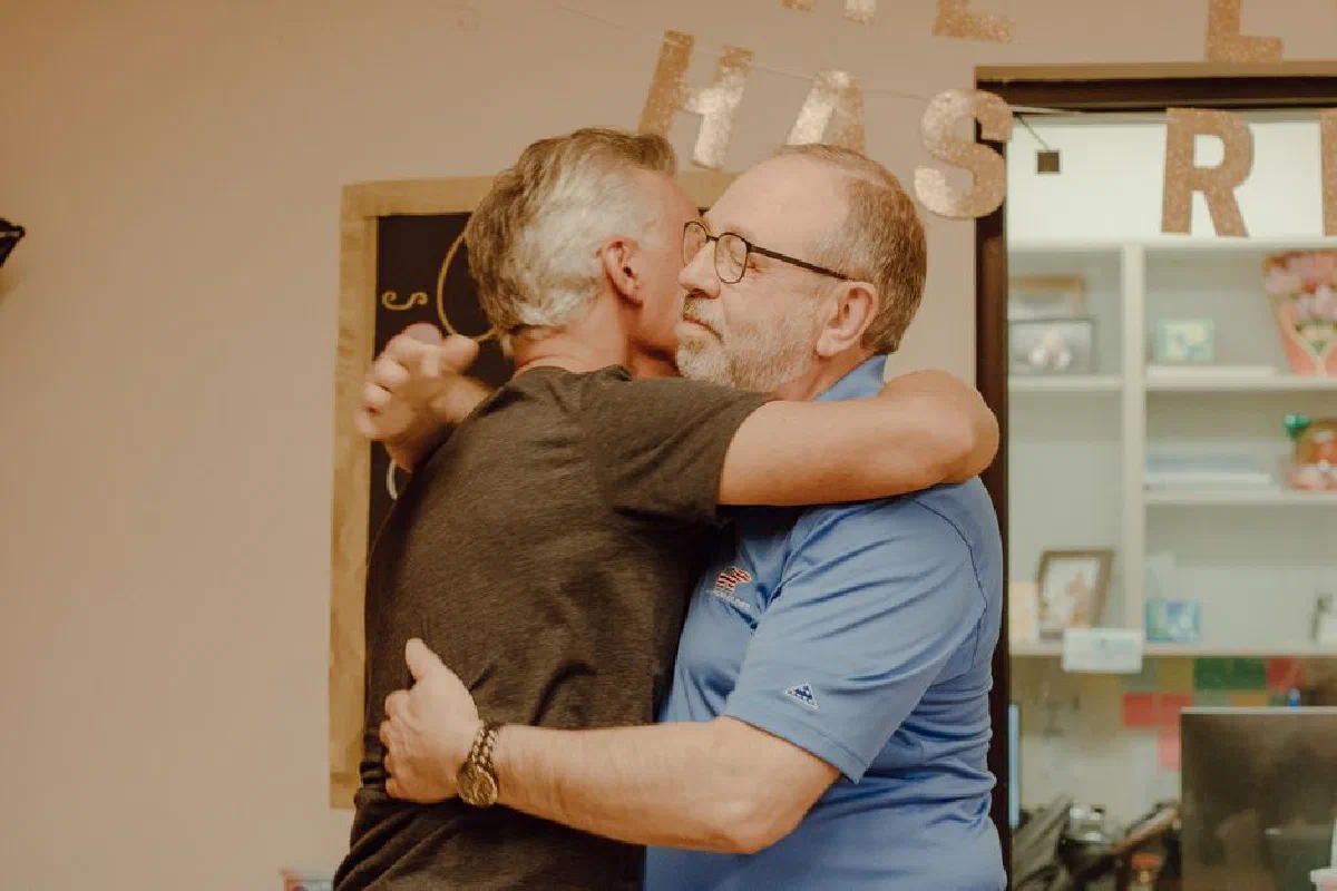 Two mature men embracing indoors, conveying friendship and affection.