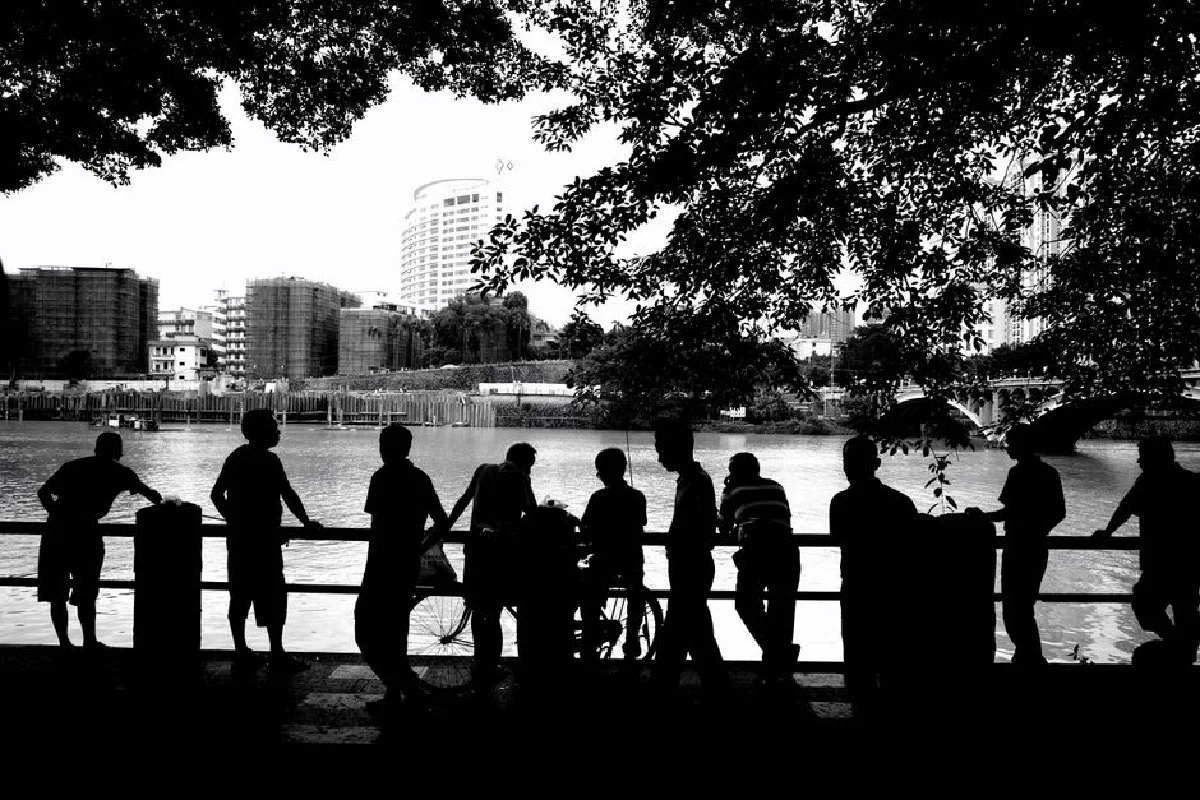 Silhouetted figures with bicycles along a riverside cityscape in black and white.