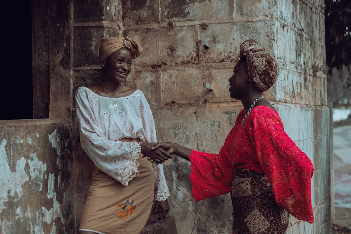 Two women in colorful traditional attire greet each other warmly by shaking hands.