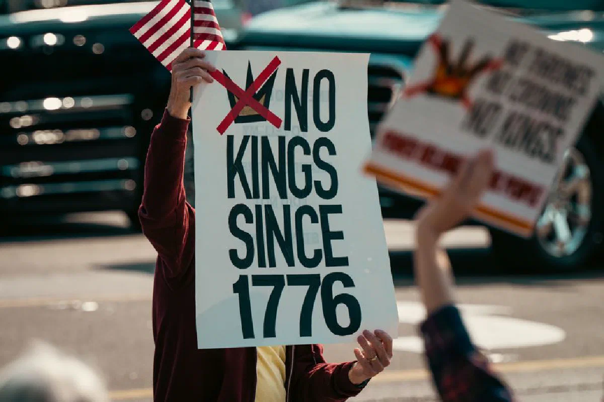 Protest sign displaying 'No Kings Since 1776' held alongside a small US flag, emphasizing democratic sentiments.