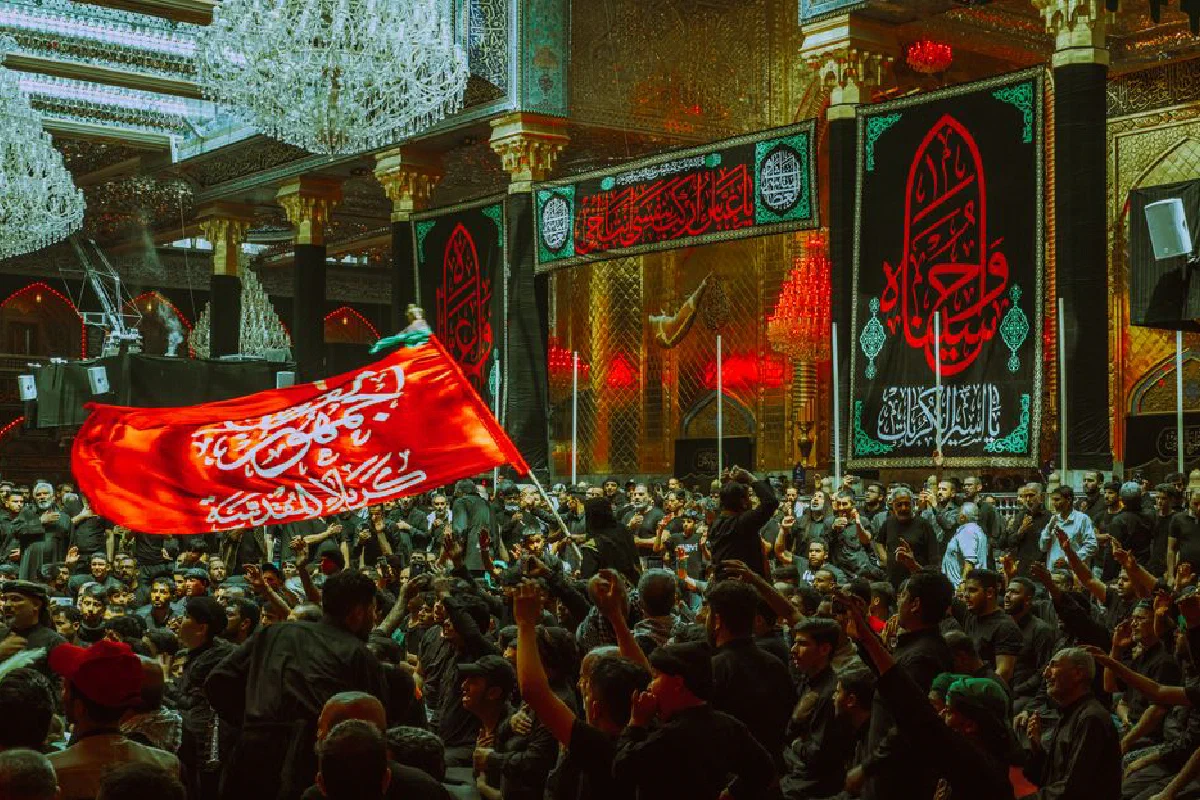 Crowd gathered in ornate mosque during a vibrant religious ceremony with red flags and decorations.