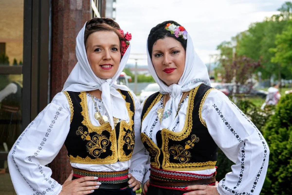 Two women in traditional Balkan costumes pose outdoors, showcasing cultural attire.