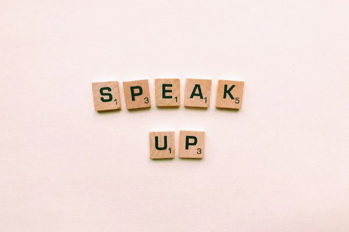Wooden Scrabble tiles spelling 'Speak Up' on a pink background.