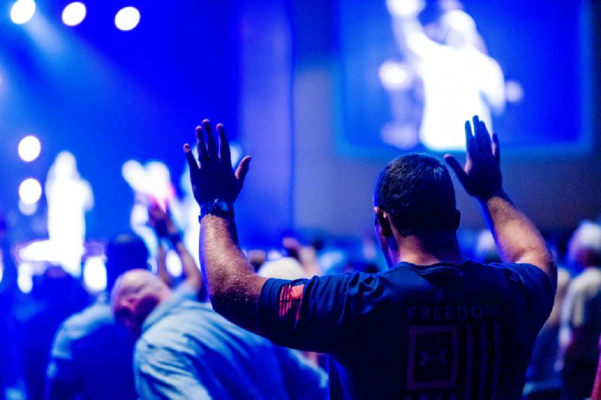 A man raises his hands in worship at a vibrant indoor event with a blurred stage.