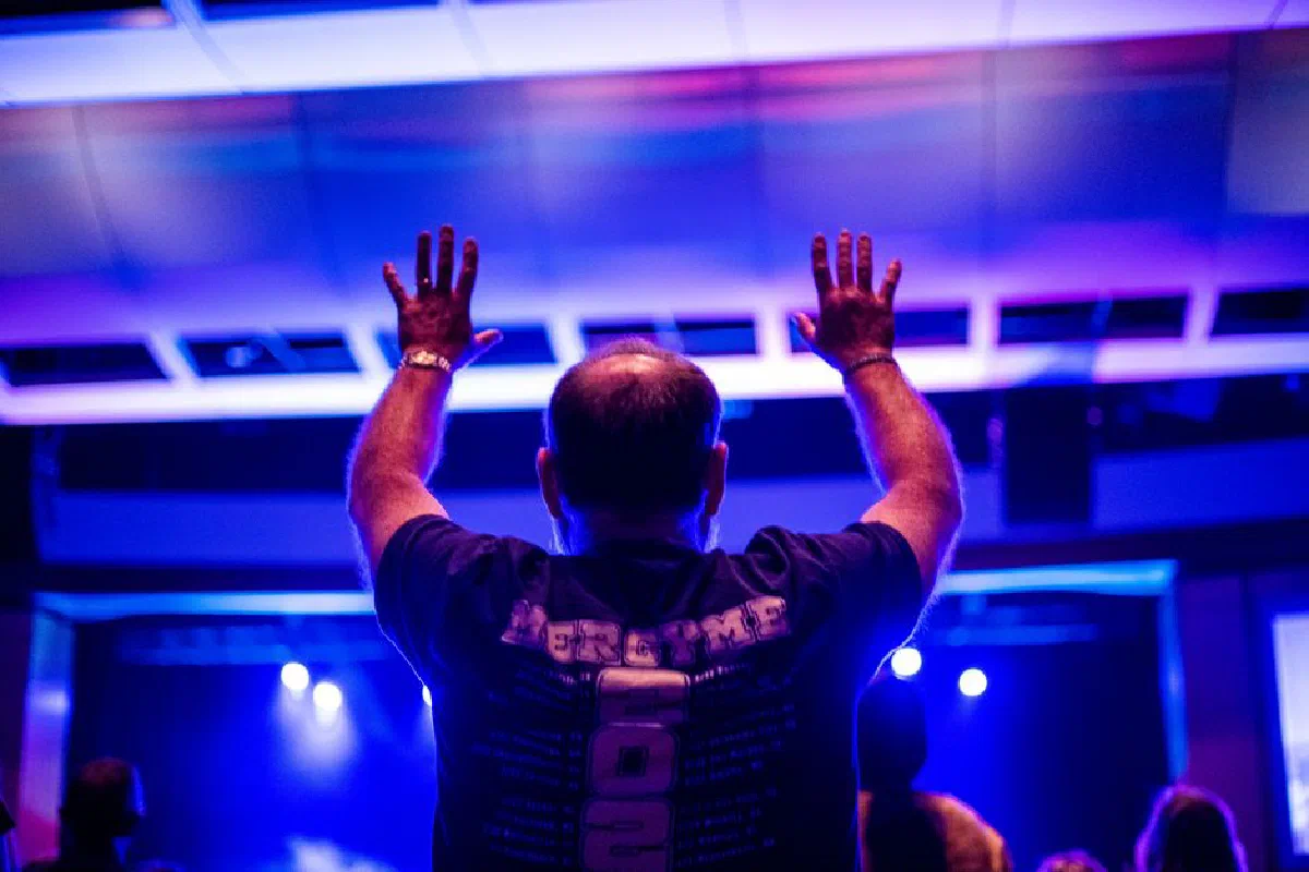 Back view of a man raising hands indoors with vibrant stage lighting at an event.