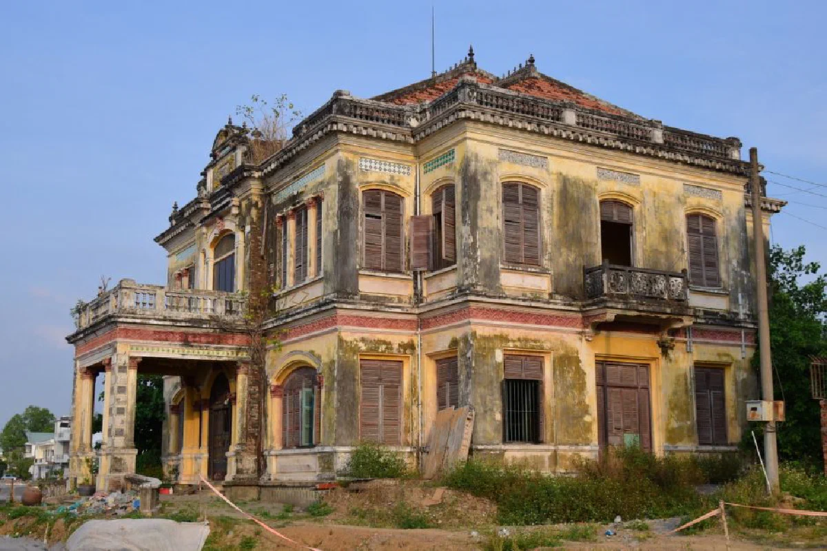 Abandoned colonial-style house with weathered facade and overgrown foliage.