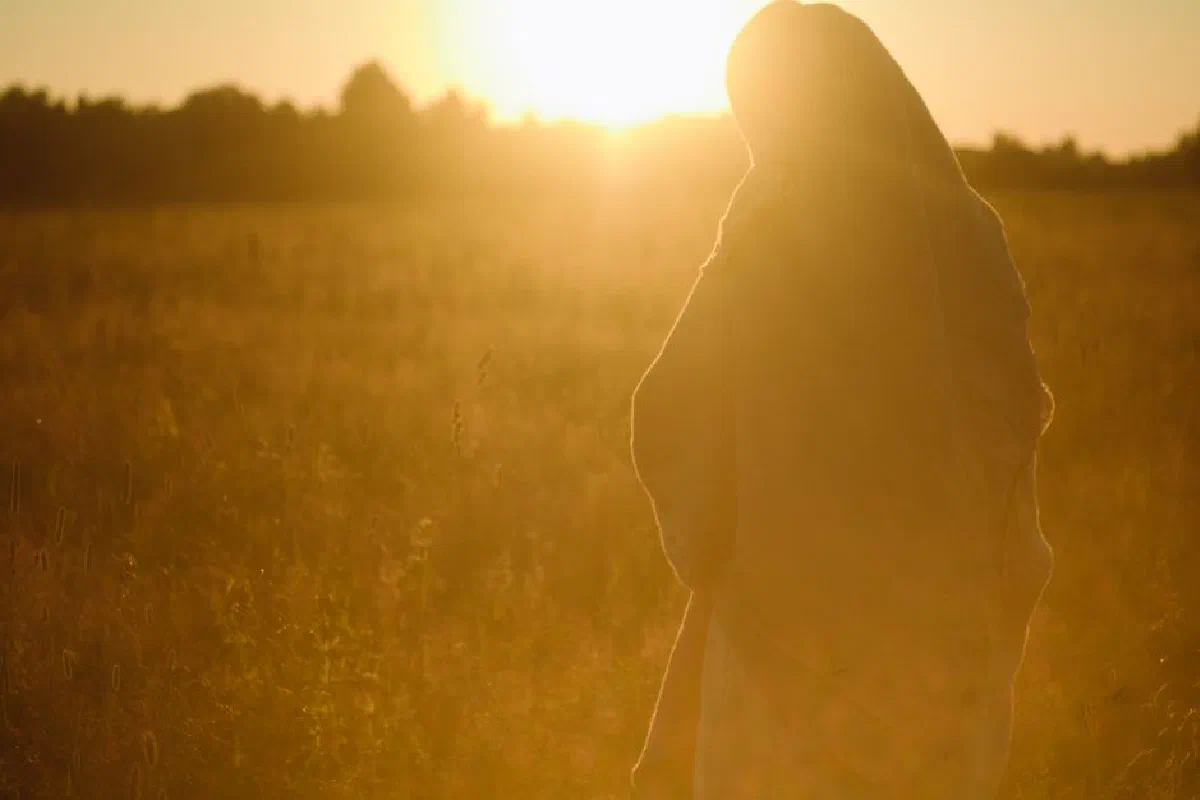 A person wrapped in a blanket standing in a sunlit field during sunset.