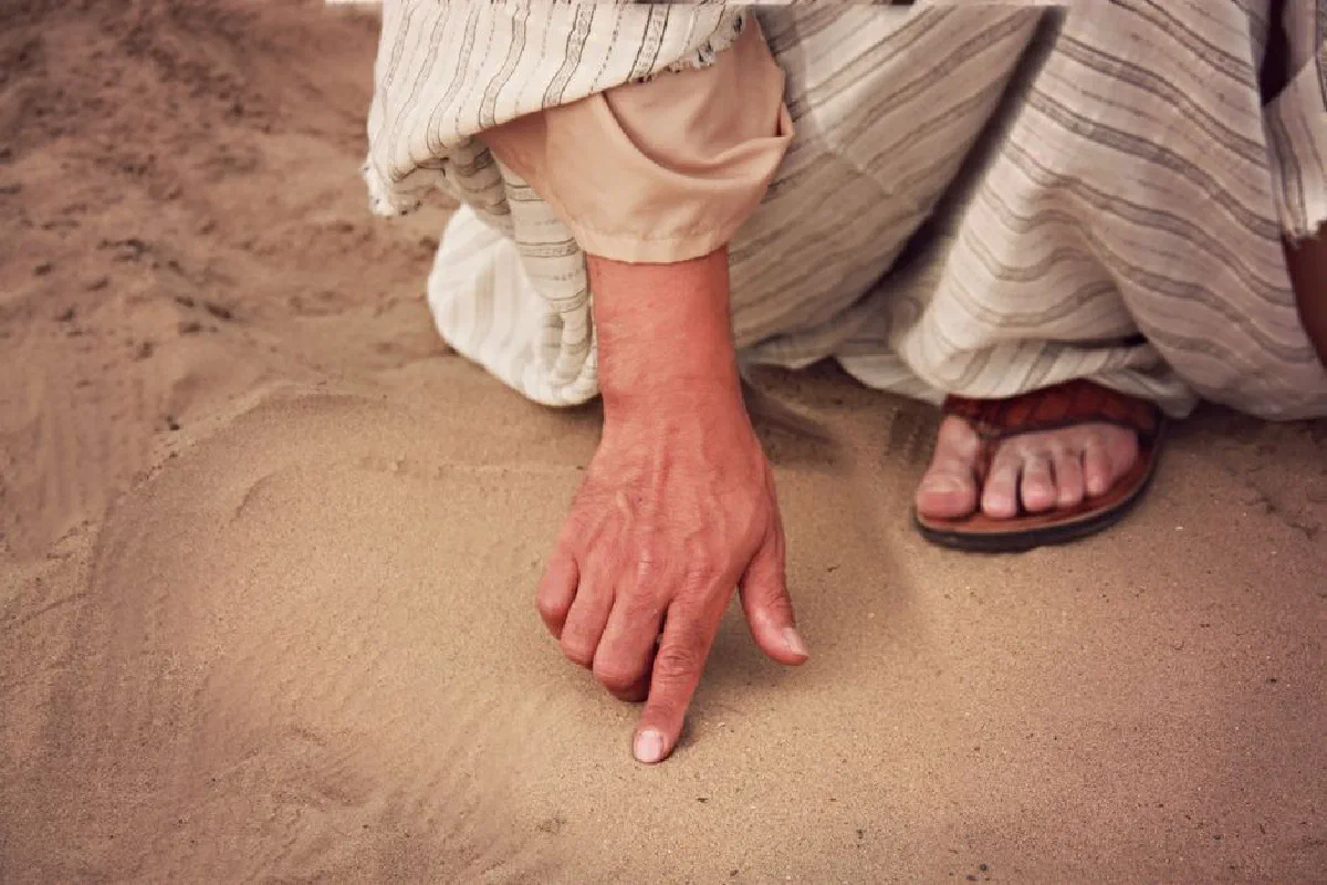 Close-up of a person drawing in sand with a finger outdoors, wearing traditional attire.