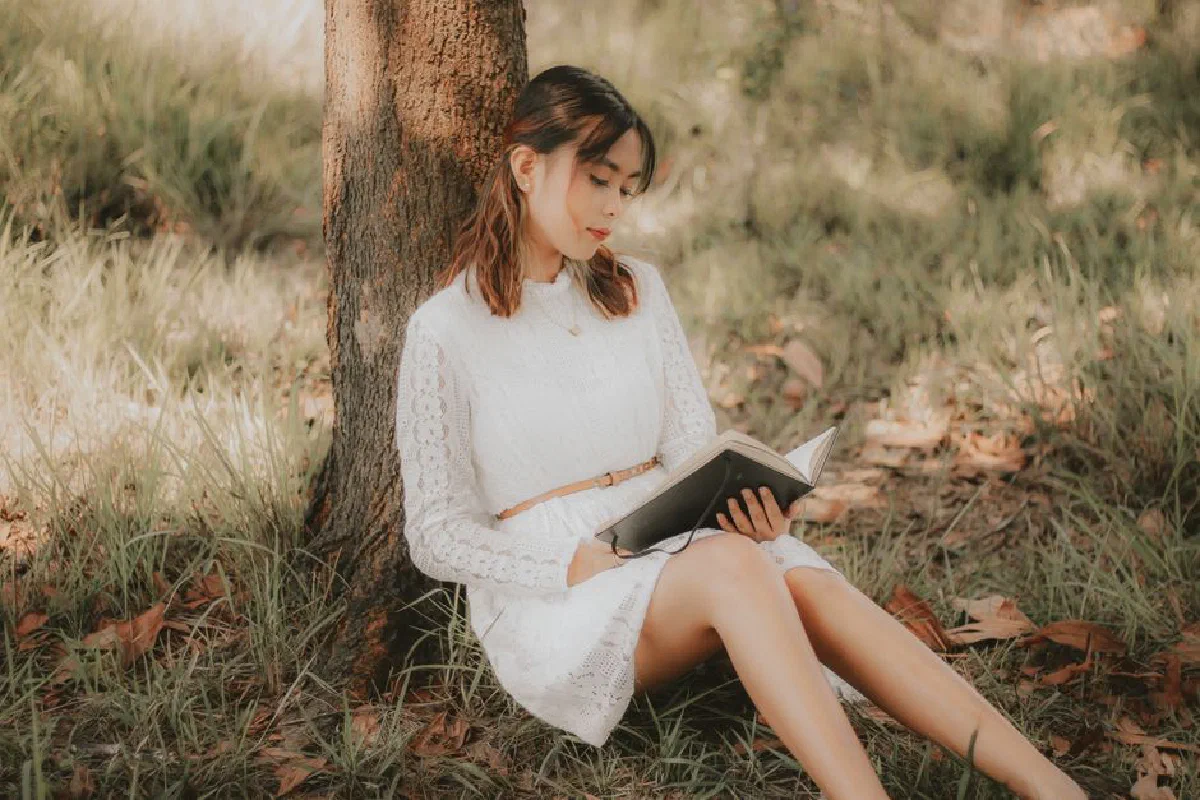 A woman in a white dress reads a book while sitting under a tree on a sunny day.
