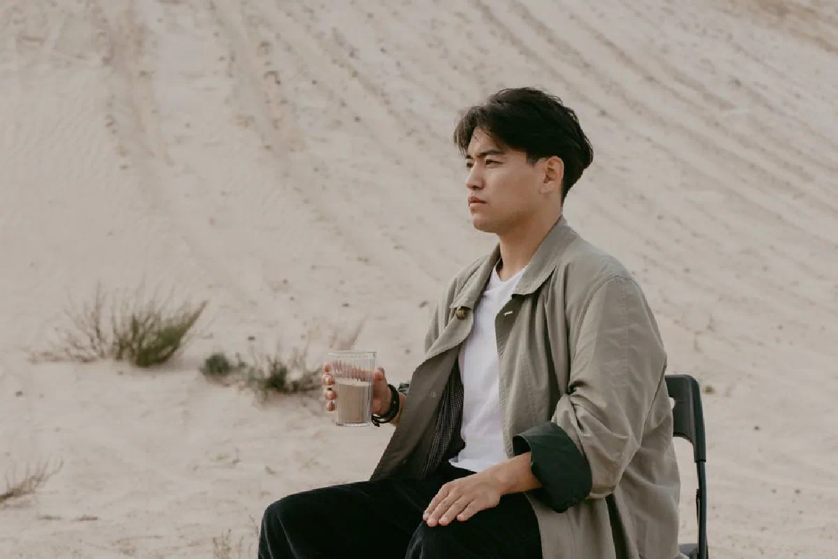 Asian man holding a glass while sitting on a chair in a sandy outdoor setting.