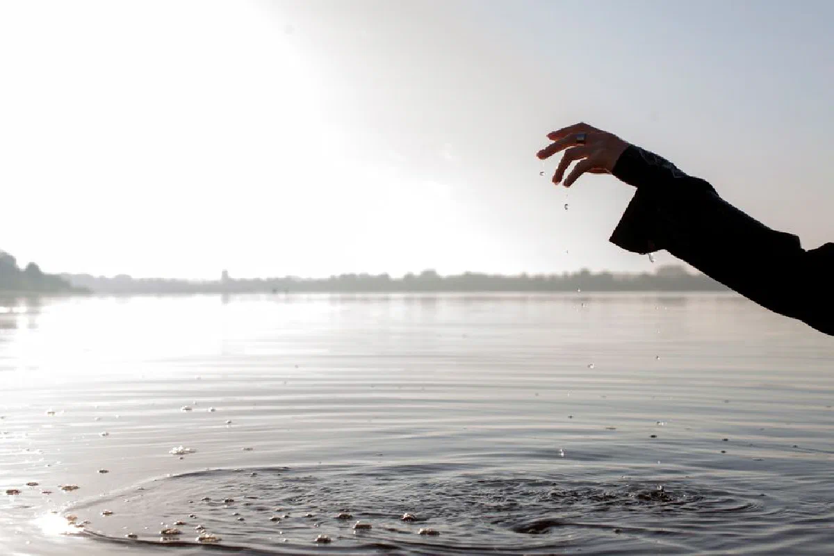 A hand captures droplets above a calm lake creating gentle ripples, evoking peace and tranquility.