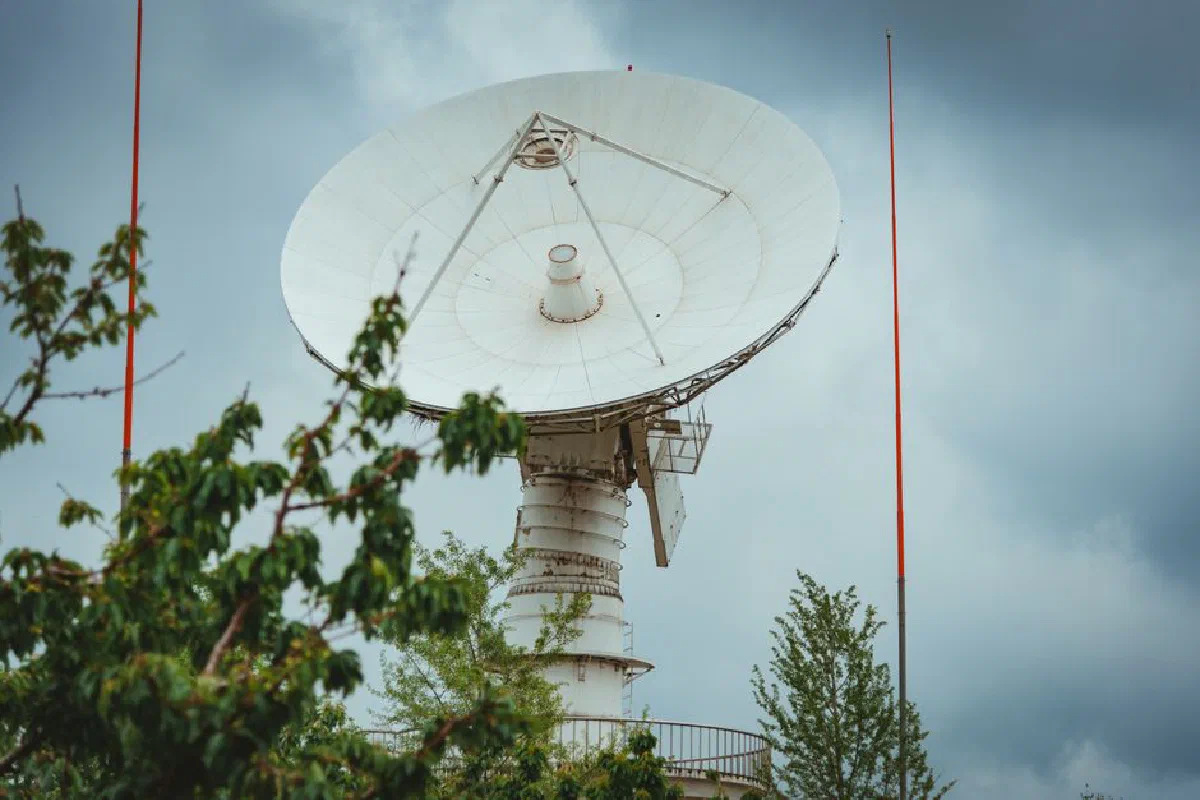 A large parabolic satellite dish with a cloudy sky background, surrounded by greenery.