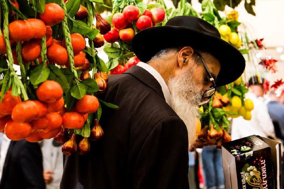 A man examines items in a vibrant market display with hanging fruit decorations.