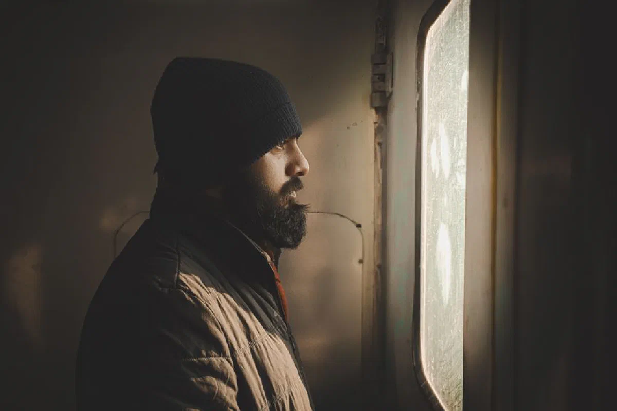 A man in a beanie and jacket looks thoughtfully out a window, illuminated by soft light.