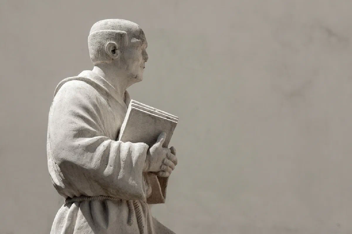 A marble statue of a monk holding a book against a plain background.