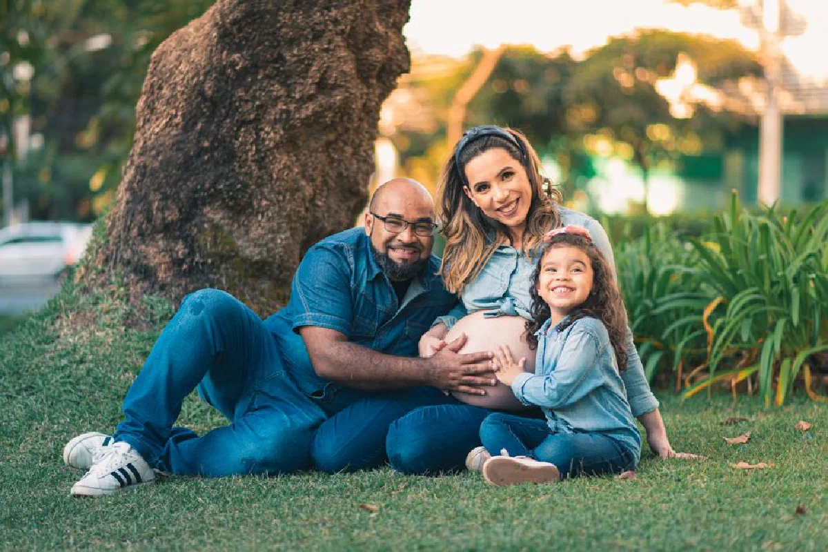 Cheerful family portrait featuring expecting mother, father, and daughter outdoors.