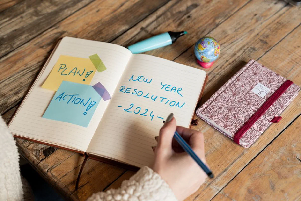 Person writing New Year 2024 resolutions in a notebook with sticky notes for planning and action.