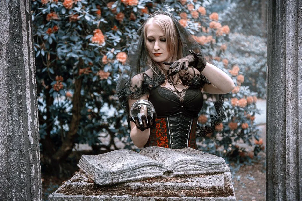A woman dressed as a witch holds a crystal ball in a mysterious outdoor setting.