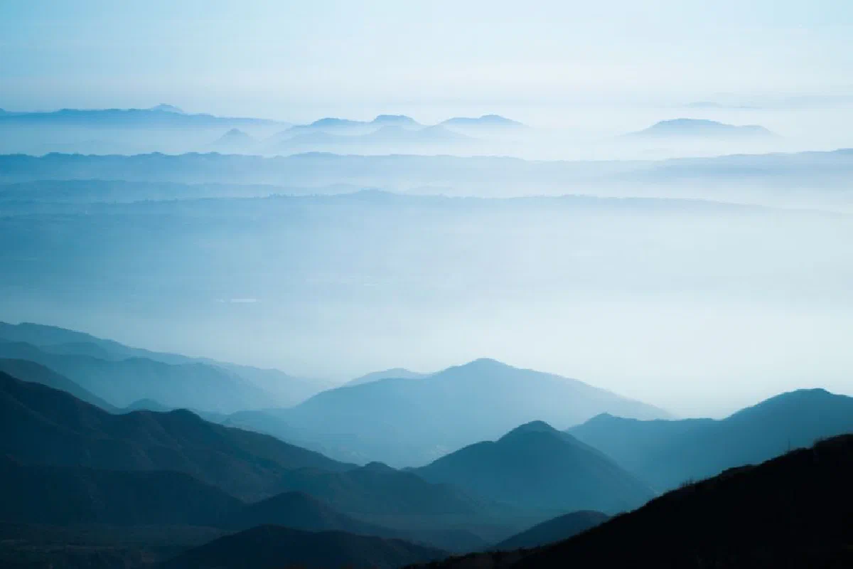 A tranquil scene of misty mountain ranges under a soft, blue sky at sunrise.