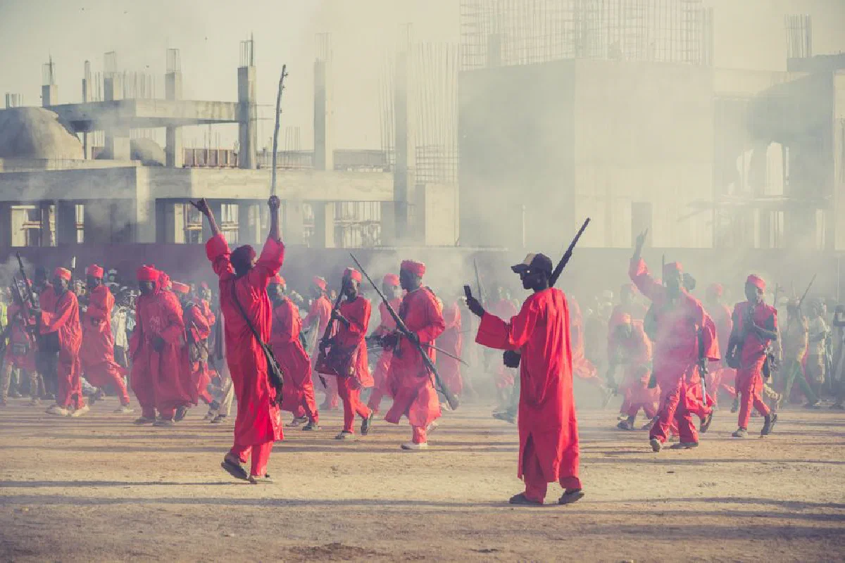 Vibrant cultural parade with people in red attire, holding sticks, amidst urban construction backdrop.