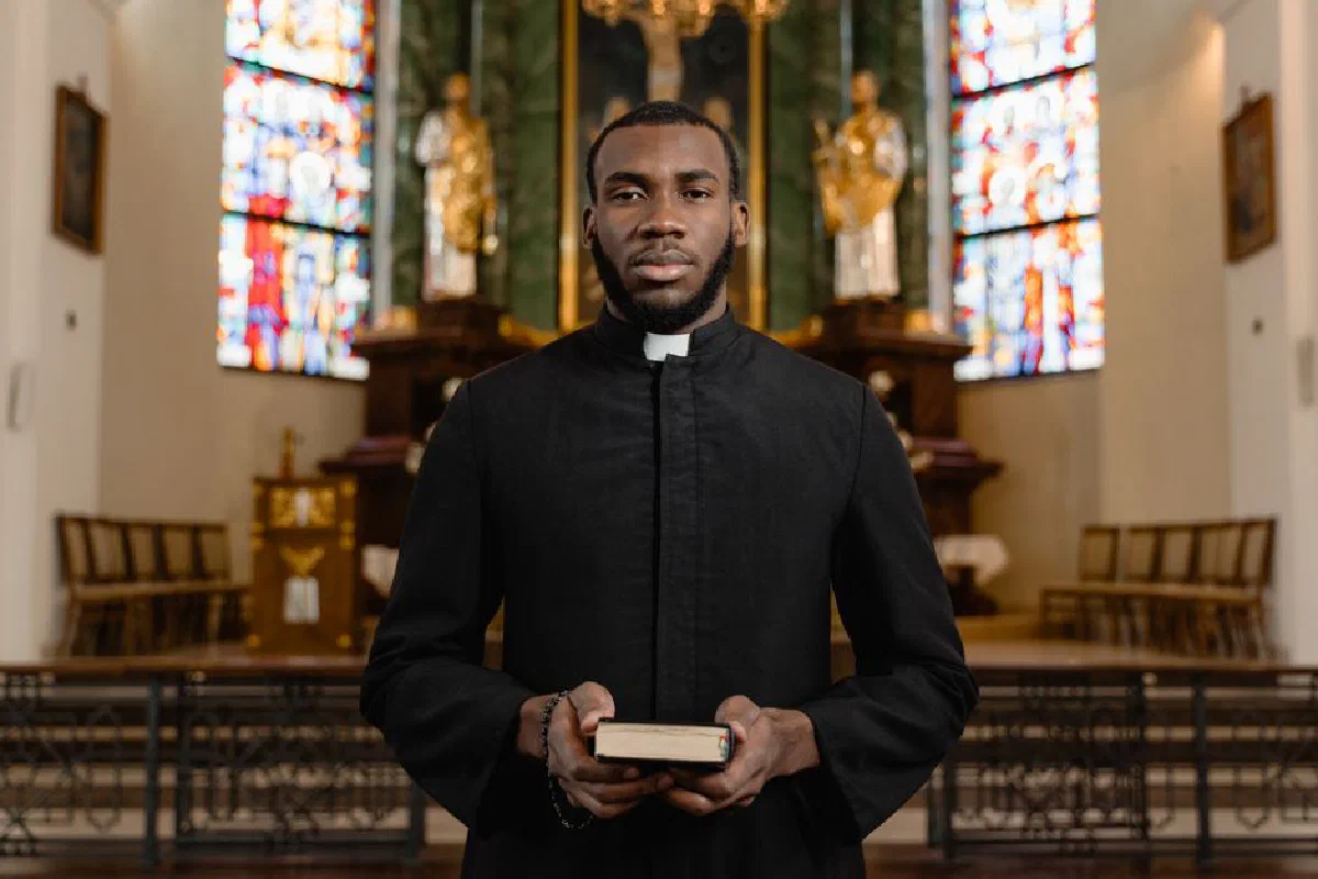 Portrait of a priest in a cassock holding a Bible in an ornate church interior.