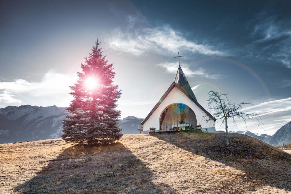 Chapel with sunburst, surrounded by mountains and tree under dramatic sky.