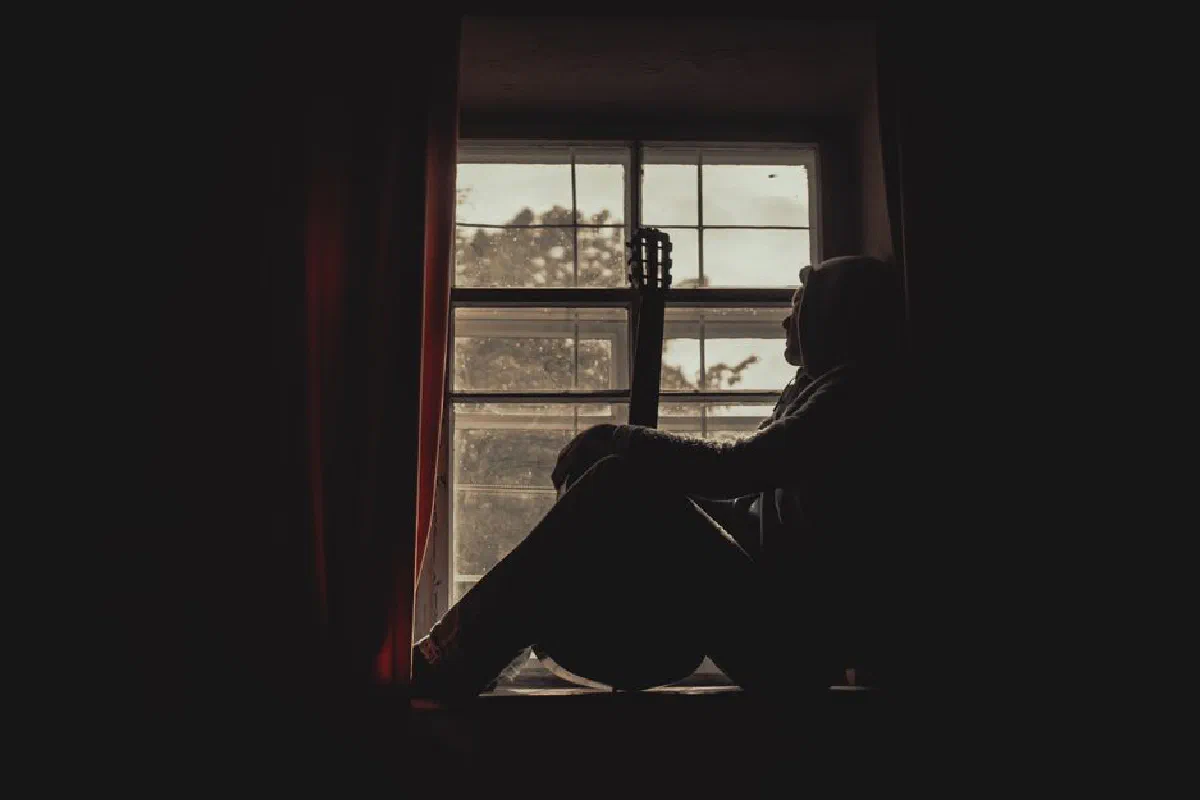 A pensive musician with guitar silhouetted by a window indoors, exuding a moody atmosphere.
