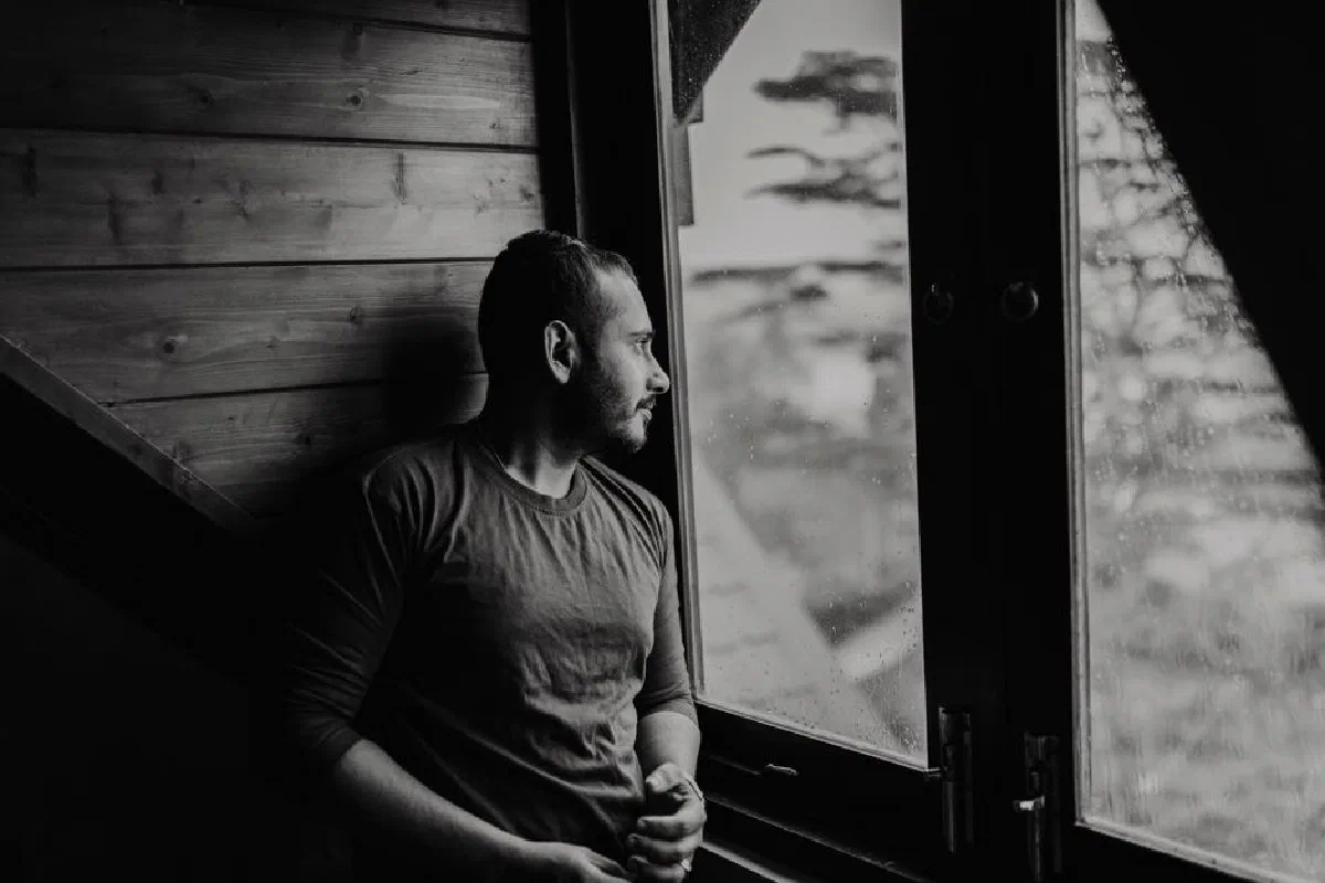 A bearded man thoughtfully gazes outside a window on a rainy day.