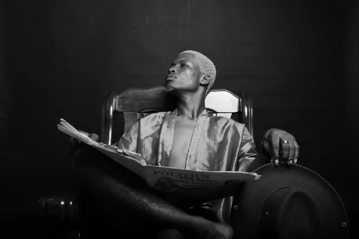 Black and white of confident African American male with dyed hair sitting in armchair and looking into distance while reading newspaper on black background