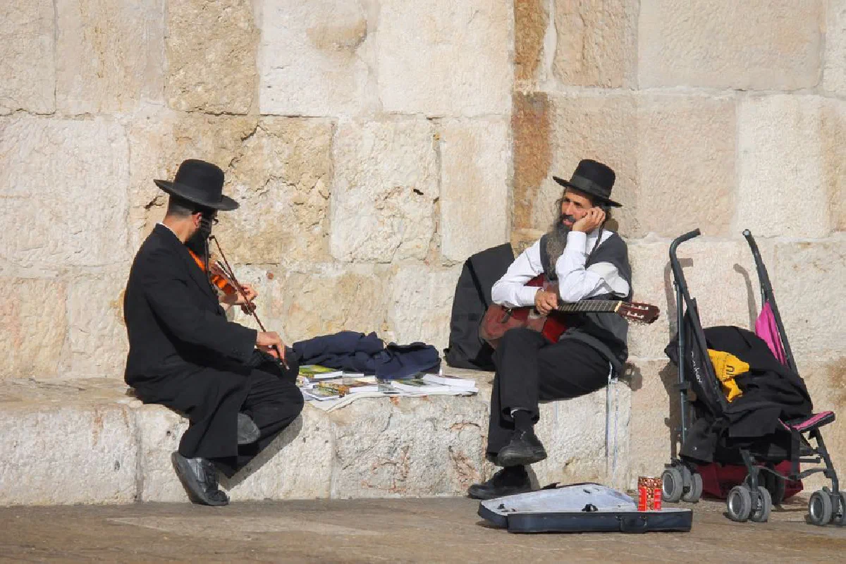Two street musicians in traditional attire playing violin and guitar near a stone wall in Jerusalem.