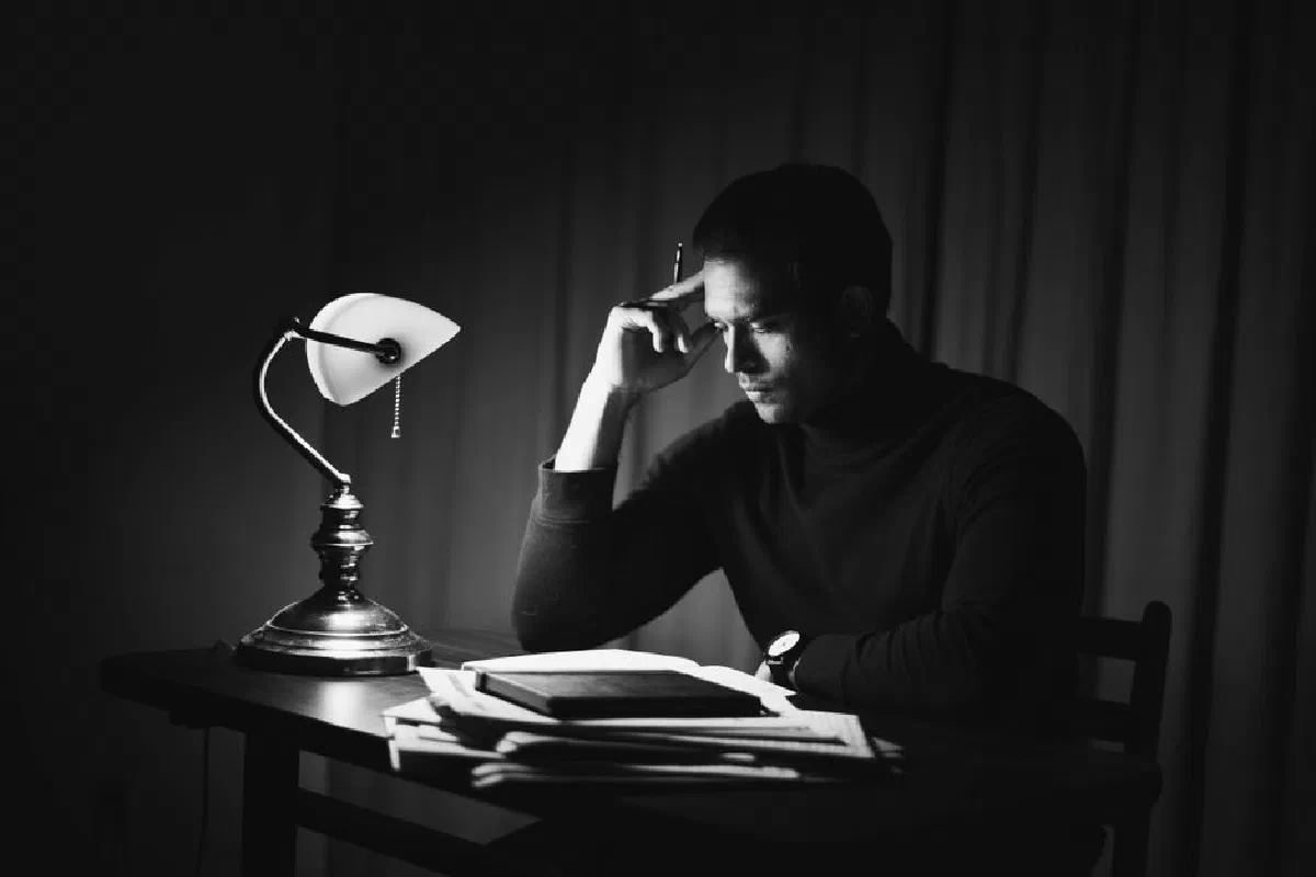 Black and white thoughtful young male in dark clothes reading book while sitting at desk with papers and bankers lamp in classy home office