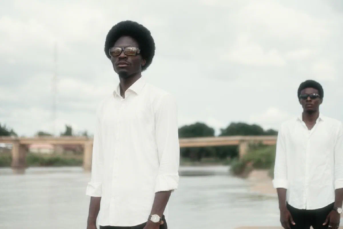 Two stylish men in white shirts and sunglasses pose by a river on a cloudy day.