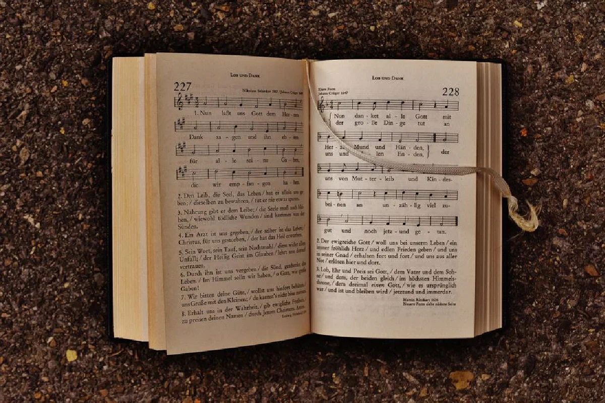 An open hymn book with musical notes laying on earthy ground, featuring sheet music and lyrics.