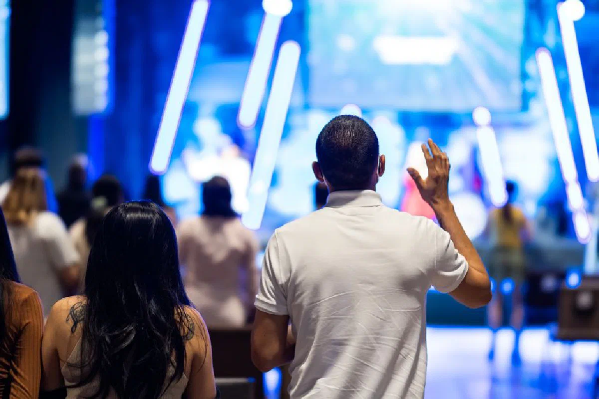 People attending a worship service indoors, featuring a man raising his hand towards the stage.