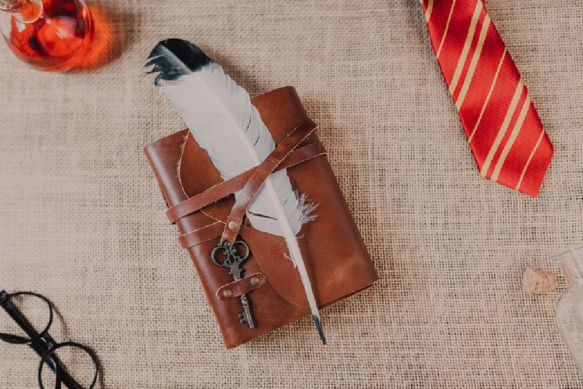 Capture of wizarding accessories including a leather spell book, feather quill, and red-striped tie on burlap.