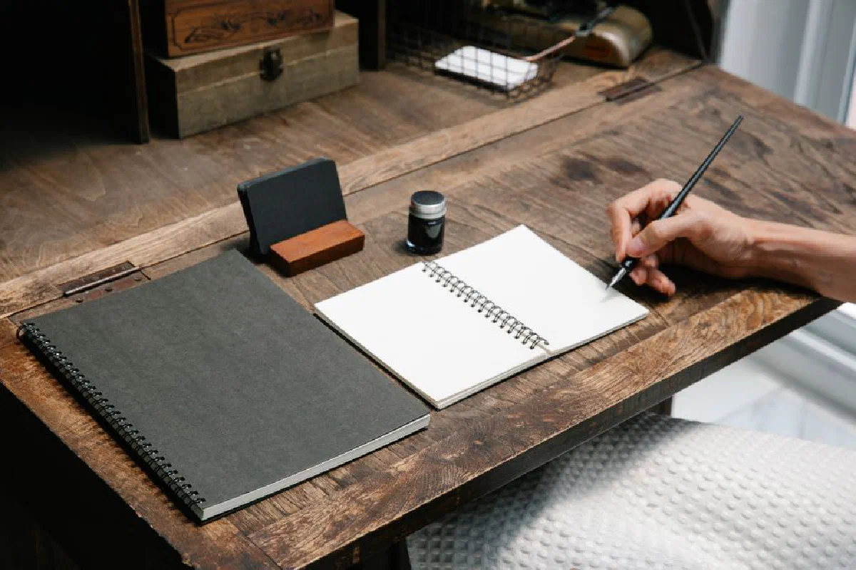 A close-up of a hand writing in a notebook at a tidy wooden desk, evoking creativity.