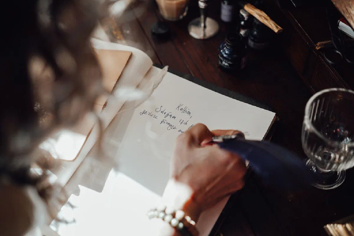 From above of crop anonymous female writer with feather taking notes in notebook at table in sunlight