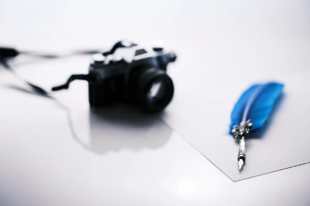 A vintage still life featuring a blurred camera and a vibrant blue quill pen on light background.