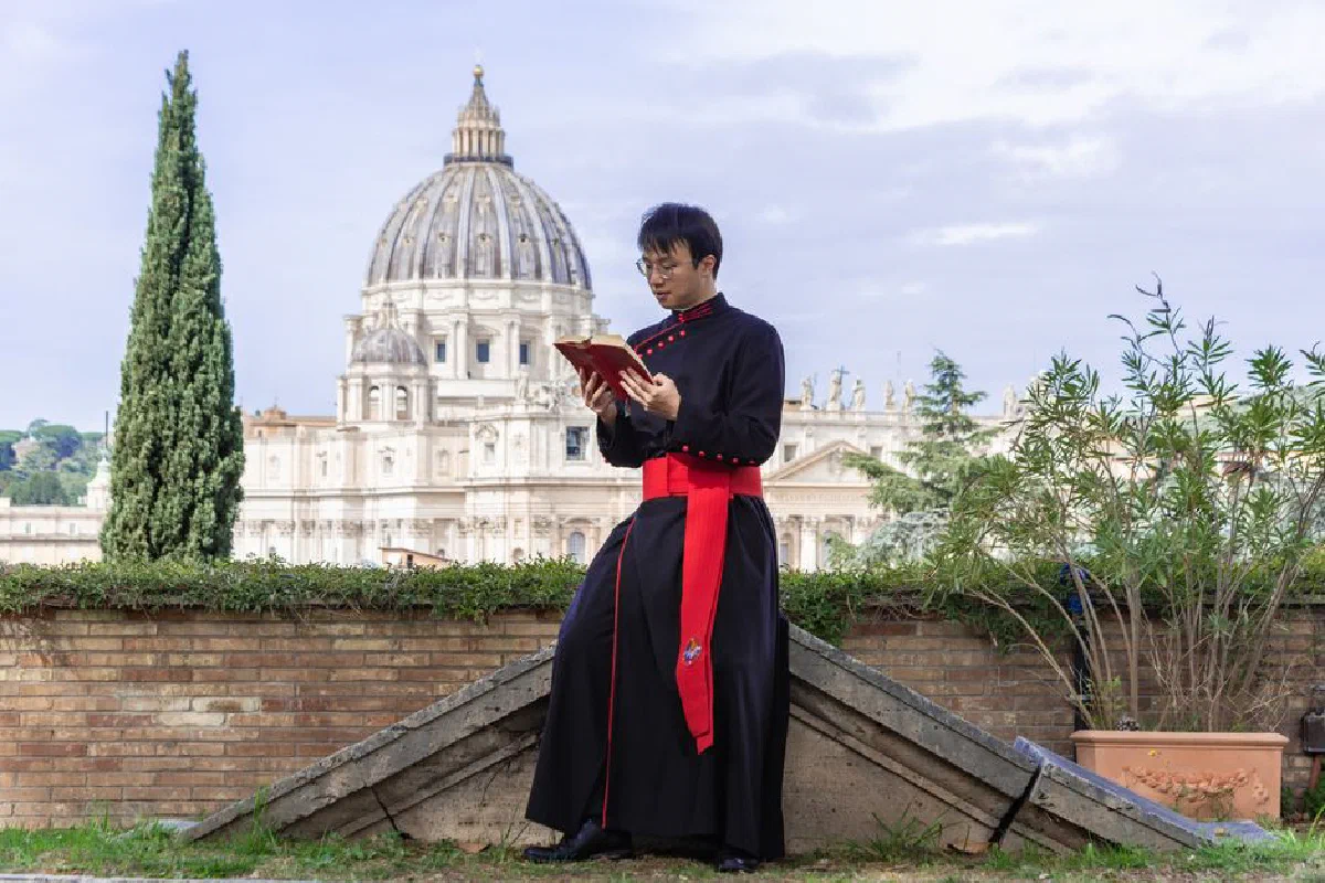 Priest in traditional attire reading a book in a garden with St. Peter's Basilica in the background.